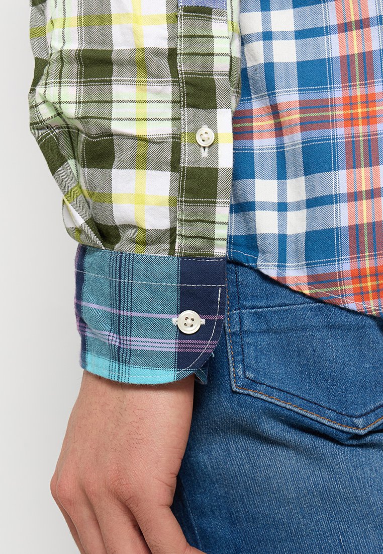 Bunt karierter Hemd mit grünen, blauen und weißen Mustern, Knopfmanschetten und einer Mischung aus Texturen. Kombiniert mit blauen Jeans.