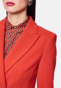 Roter, strukturierter Blazer mit markanten Revers, darüber ein rot-schwarz gemustertes Hemd. Auffälliges großes silbernes Ohrring-Accessoire.