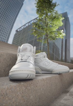 Paar weiße New Balance High-Top-Sneaker auf Außentreppen aus Beton platziert, mit modernen Glasgebäuden und einem Baum im Hintergrund.