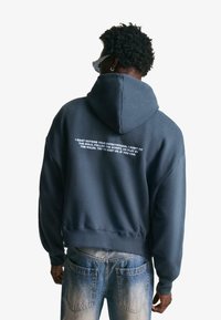 Man with short curly hair wears dark blue hooded sweatshirt with white text and light washed jeans, standing facing away.