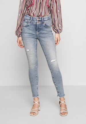 Frau trägt hellblaue, ausgewaschene Skinny-Jeans mit ausgefranstem Saum, kombiniert mit weißen Riemchen-High-Heels und einer langärmeligen gemusterten Bluse.