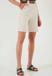 Pantaloni corti beige chiaro con vita alta, orli arrotolati e tasche laterali. Abbinati a una cintura marrone e sandali a strisce verde scuro.