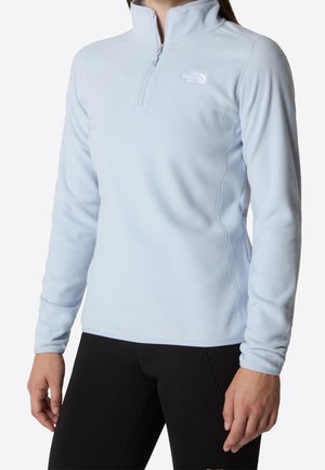 Femme portant un pull polaire bleu clair à manches longues avec une fermeture éclair au quart et un legging noir, debout devant un fond blanc.