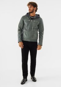 Giacca di pelle verde con dettagli trapuntati sulle spalle e cerniere frontali, indossata sopra una felpa con cappuccio grigia. Pantaloni neri e stivali completano l'outfit.