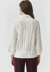 Maglione lavorato a maglia color crema con collo alto, design a trecce e maniche ampie. I polsini e l'orlo a coste aggiungono un dettaglio di texture.