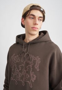 Felpa con cappuccio marrone con un logo grafico che raffigura tre leoni delineati. Accompagnata da un berretto beige, il tessuto ha una texture liscia.