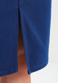 Blå denimkjol med en sidofålla, rak skärning och kontrasterande trådstickning vid fåll och slitskant. Slät textur.