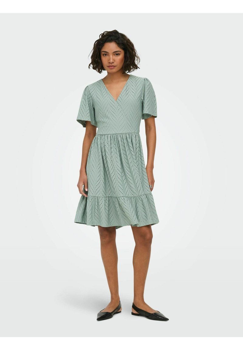 Femme debout portant une robe vert clair jusqu'aux genoux avec de courtes manches flottantes et un motif chevron à pois subtil, associée à des ballerines noires.
