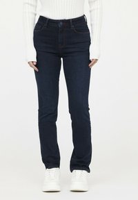 Jeans de color azul oscuro con cintura alta, ajustados, que cuentan con un cierre de botón y costuras rojas en contraste. Combinados con zapatillas blancas.