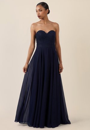 Frau trägt ein trägerloses, bodenlanges Marineblaues Kleid mit Spitzenoberteil und fließendem Rock, steht vor einem schlichten beigen Hintergrund.