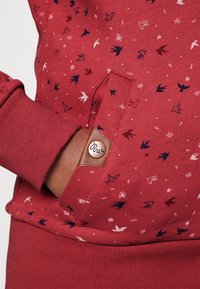 Hånd i lommen på en rød sweatshirt med små fugle- og blomster mønstre, manchetærmer, og en brun læderlap med knapdetalje.