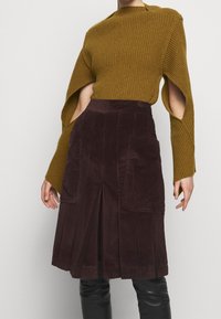 Oliivinvihreä ribatattu neuletakki, jossa on leikattu hihat, yhdistettynä tumman viininpunaisiin farkkuhameeseen, jossa on kaksi etutaskua ja pleiattuja yksityiskohtia.