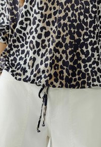 Leopardenmuster-Oberteil in Schwarz-, Beige- und Cremetönen, mit lockerer Passform und verstellbarem Kordelzug am Saum, kombiniert mit weißen Hosen.
