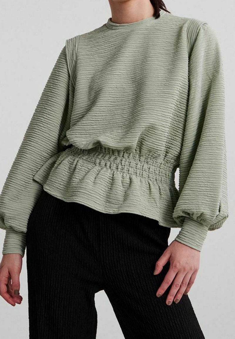 Femme portant un haut texturé vert clair à manches longues avec taille et poignets froncés, associé à un pantalon texturé noir, debout devant un fond uni.