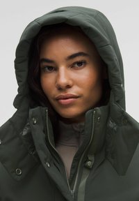 Zelená bunda s kapucí s texturovaným povrchem, patentkami a zipy. Vnitřní vrstva je v jasnější barvě, viditelná na límci a rukávech.