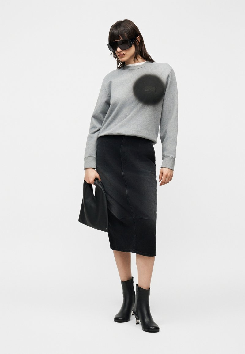 Donna che indossa occhiali da sole neri oversize, felpa grigia, gonna midi nera, stivaletti neri, che tiene una borsa nera su sfondo bianco.