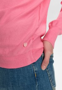 Maglione rosa lavorato a maglia con texture a costine, caratterizzato da un piccolo dettaglio con bottone rosa sul lato. Abbinato a jeans in denim blu.