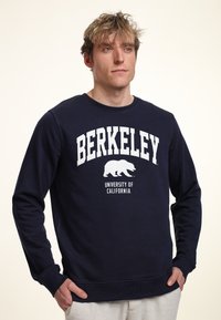 Felpa blu navy in cotone, con collo a girocollo, polsini a coste e un design con la scritta "BERKELEY" e un grafiche di un orso in bianco.