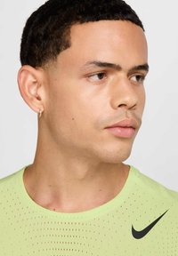 T-shirt de sport vert clair avec une texture perforée et un logo Nike noir. Gros plan sur le profil d'un jeune homme, présentant des cheveux courts et bouclés ainsi qu'une boucle d'oreille.