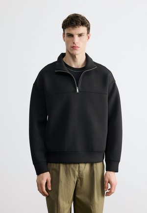 Νέος άντρας που φοράει μαύρο πουλόβερ με φερμουάρ στο λαιμό και χακί παντελόνι, στέκεται μπροστά από ένα απλό φωτεινό γκρι φόντο.