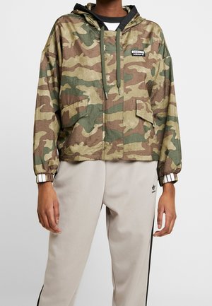 Person trägt eine grün-braune Camouflage-Kapuzenjacke mit Vordertaschen und hellbeige Adidas Trainingshose mit schwarzen Streifen.