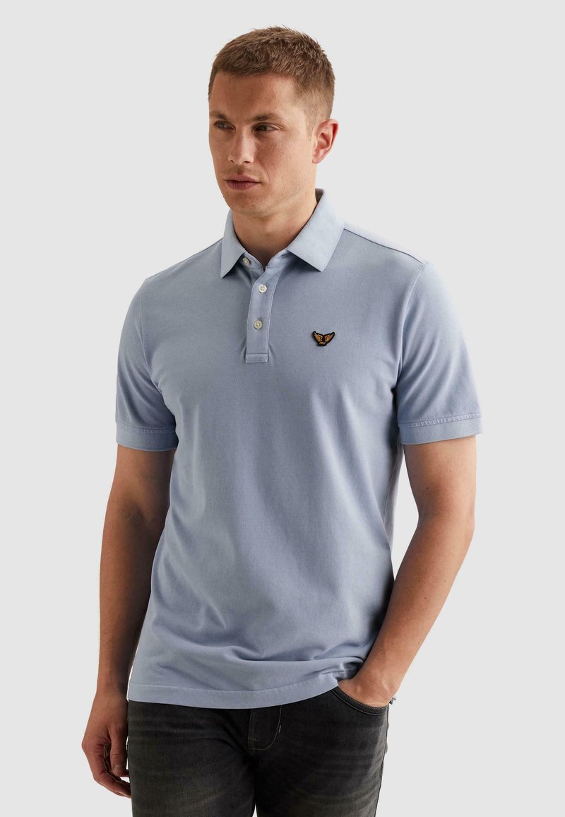 Homme portant un polo bleu clair à manches courtes avec un petit logo brodé sur la poitrine gauche, associé à un pantalon foncé, debout, une main dans la poche.