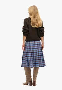 Pull marron tricoté avec manches retroussées, associé à une jupe midi à carreaux bleue avec des accents marron et jaune, et des bottes grises montantes jusqu'aux genoux.