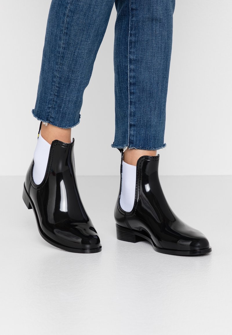 Personne portant des bottines de pluie noires brillantes avec des panneaux élastiques blancs sur les côtés et un jean bleu court avec des ourlets effilochés.