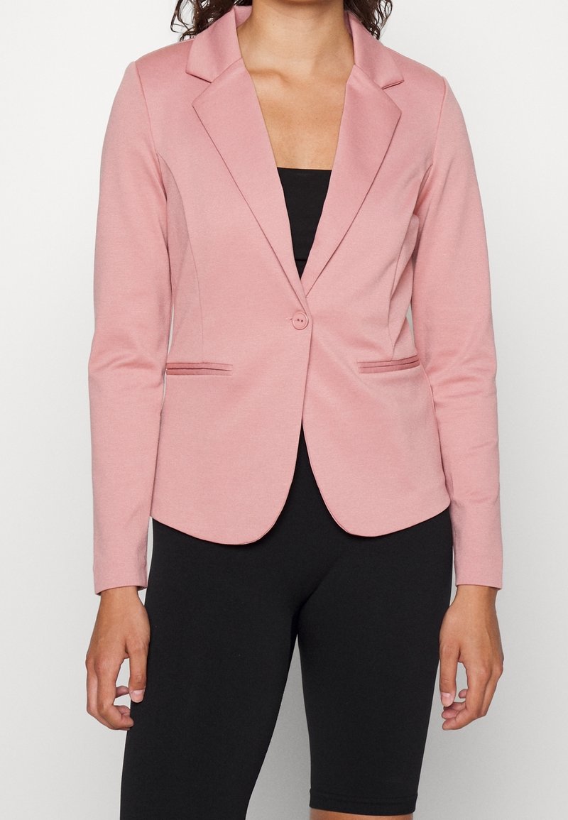 Femme portant un blazer rose ajusté par-dessus un top noir et des leggings noirs jusqu’aux genoux, debout, les bras détendus le long du corps.