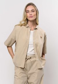 Beige kortärmad skjorta i lätt material med knäppning fram och ett litet blommönster broderat på bröstet.