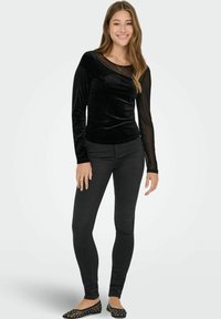 Mladá žena se usmívá, má na sobě černý sametový vršek s průsvitnými dlouhými rukávy, černé skinny džíny a černé ploché boty s cvočky, stojí na bílém pozadí.