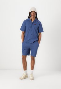 Chemise bleue à manches courtes avec col et fermeture à boutons, associée à un short bleu assorti. Comprend un petit logo. Les chaussures sont beiges avec des lacets.