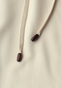 Tissu beige avec une couture double et deux cordons crème se terminant par des perles en bois brun foncé.