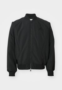 Veste bomber noire Adidas avec fermeture éclair à l'avant, col, poignets et ourlet côtelés, et petite poche zippée sur la manche gauche.