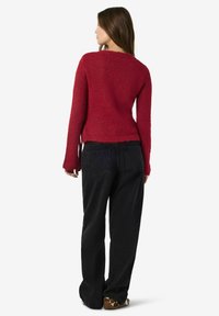 Kvinna med långt brunt hår, iklädd en röd stickad tröja, svarta lösa jeans och mönstrade skor, stående vänd bort mot en vit bakgrund.