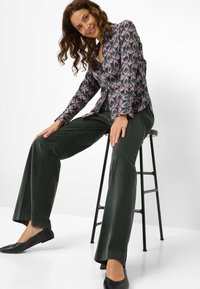Chemise à motif avec un col montant et des manches longues, associée à un pantalon large vert foncé et des chaussures plates noires, assise sur un tabouret noir.
