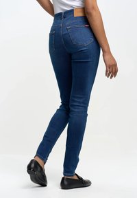 Inimene, kes kannab kitsaid tumesiniseid teksapükse ja musti mokasiine, kõndides ühe jalaga kergelt tõstetud, tavalise valge tausta ees.