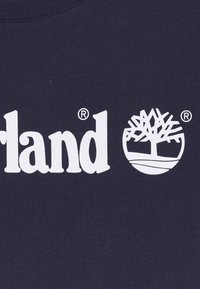 Camiseta de algodón azul marino oscuro con texto de logo impreso en blanco "Timberland" y un gráfico estilizado de árbol debajo. Mezcla de tela texturizada.