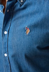 Camisa de mezclilla azul con cuello abotonado, botones blancos y un logo bordado de un jinete en rojo y dorado.