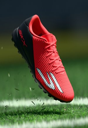 Roter und schwarzer Fußballschuh mit weißem Logo auf grünem Gras mit weißer Linie, zeigt Vorder- und Seitendetails sowie strukturierte Oberfläche.