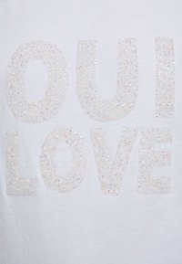 Bílá tričko s velkými, třpytivými písmeny nápisem "OUI LOVE", s texturovaným, třpytivým povrchem a hladkým tkaninovým pozadím.