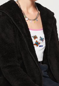 Zwarte, fuzzy hooded jacket over een witte tanktop met kleurrijke geborduurde vlinders en de tekst "OBEY", aangevuld met een zilveren ketting.