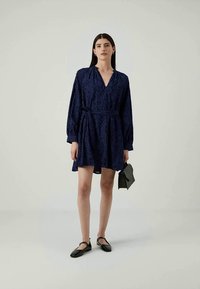 Morsko plava haljina s V-izrezom, dugim rukavima i floralnom teksturom. Uključuje remen oko struka te se nosi s crnim balerinkama i torbom.