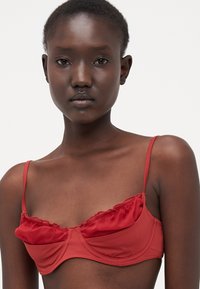 Reggiseno in satin rosso con ferretto. Presenta un bordo arricciato lungo la parte superiore e spalline sottili e regolabili. Texture liscia, design strutturato.
