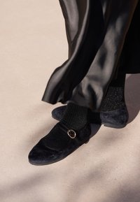 Czarne płaskie buty zamszowe z okrągłą sprzączką, noszone z czarnymi teksturowanymi skarpetkami, pod zwiewną czarną tkaniną.