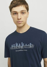 Jūros mėlynos spalvos medvilninė marškinė, puošta dideliu baltu tekstu "JACK & JONES" ir miesto panorama. Su apvaliu iškirpimu ir trumpomis rankovėmis, lygi tekstūra.