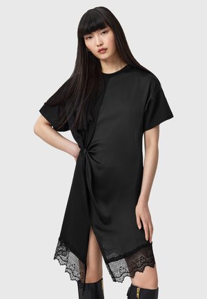 Black shirt dress with a twisted side detail, asymmetrical hem, and lace trim. Made of smooth fabric, featuring short sleeves and a side slit.