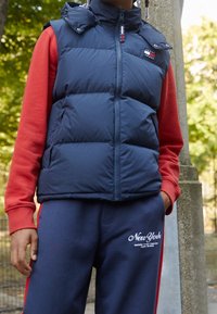 Granatowa pikowana kamizelka z wysokim kołnierzem i zamkiem błyskawicznym, zestawiona z czerwoną koszulą z długim rękawem oraz szarymi spodniami dresowymi z białymi akcentami tekstowymi.