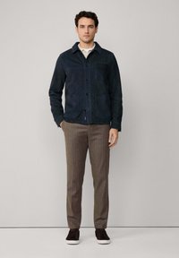 Marineblauwe suède jas met knoopsluiting, twee voorkruidge zakken. Gecombineerd met bruine gestreepte broek en donkere sneakers, op een neutrale achtergrond.