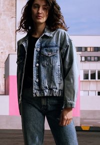 Jaca de ganga com um design encurtado, lavagem azul clara, frente com botões, dois bolsos no peito e bainha elástica. Usada com jeans de cintura alta a condizer.